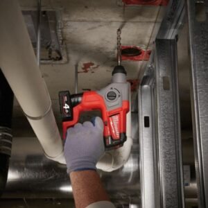 Thợ đeo găng dùng máy khoan bê tông Milwaukee M12 CH-0C khoan ngược lên trần, cạnh đường ống trắng và giá treo kim loại