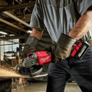 Thợ đeo găng da cổ dài cầm máy mài pin Milwaukee M18 mài chi tiết thép, tia lửa bắn ra, phía sau là nhà xưởng kèo thép