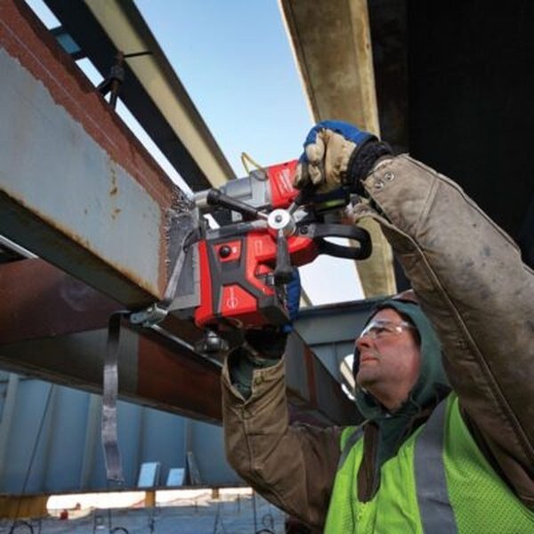 Thợ đội mũ trùm, đeo kính và áo phản quang dùng máy khoan từ Milwaukee M18 khoan ngược lên dầm thép ngoài công trình