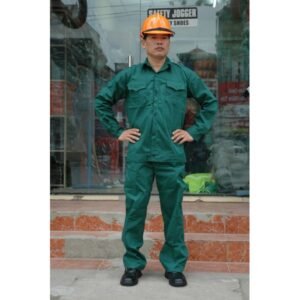 Nam công nhân mặc bộ quần áo bảo hộ kaki màu xanh két đậm, áo dài tay hai túi nắp, đội mũ bảo hộ cam, đứng chống hông